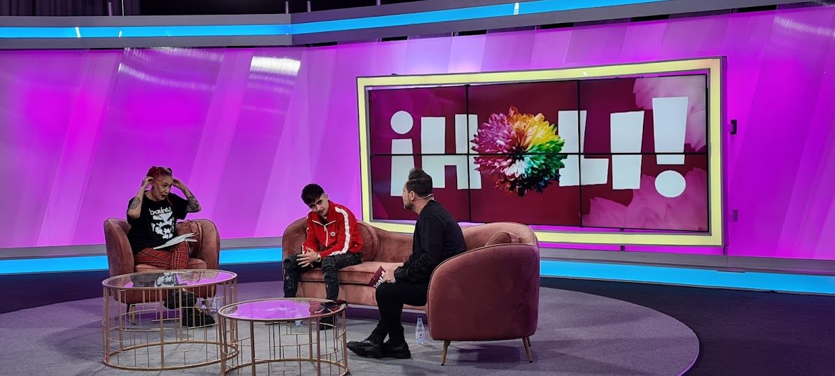 Un colorido plató de televisión con dos presentadores y un invitado sentados en un sofá rosa.