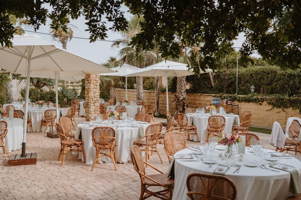 Espacio para eventos al aire libre con mesas redondas, sillas de mimbre y sombrillas rodeado de palmeras en 1180studio Wedding Planner.