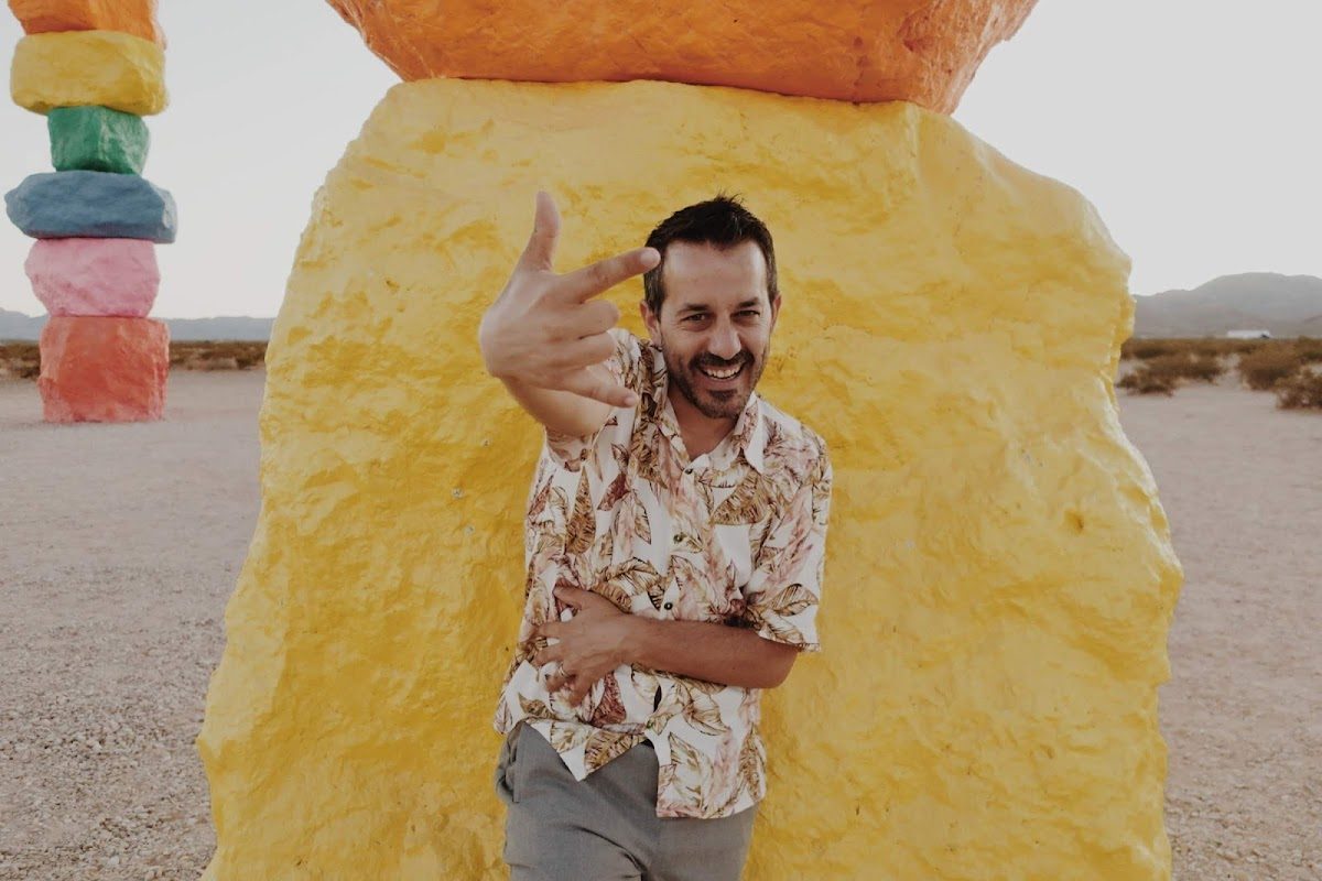 Un hombre con una camisa estampada de flores posa de forma divertida frente a una gran formación rocosa de colores.