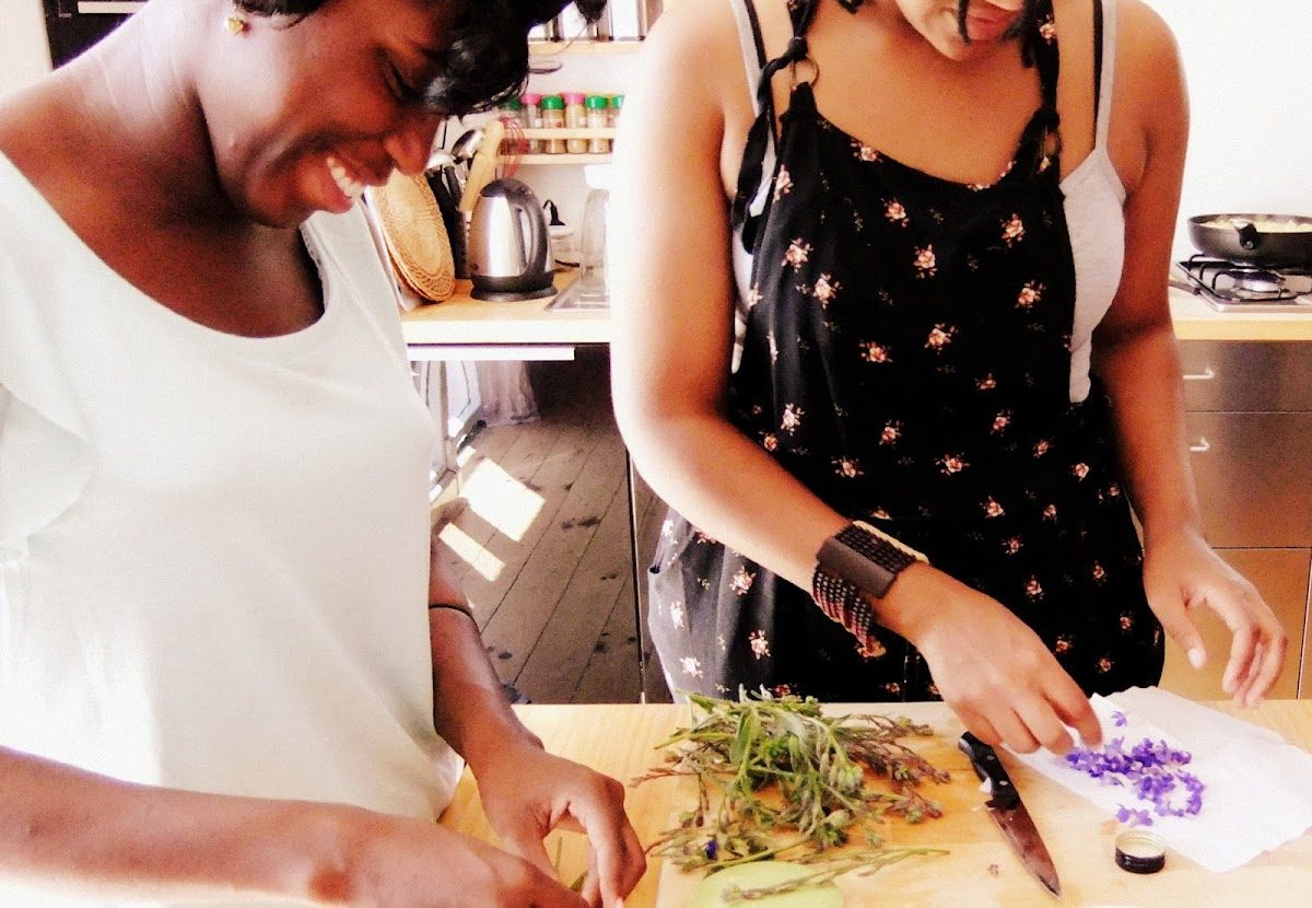 Two women preparing herbs and flowers on a cutting board in a bright kitchen at A Cooking Day.
