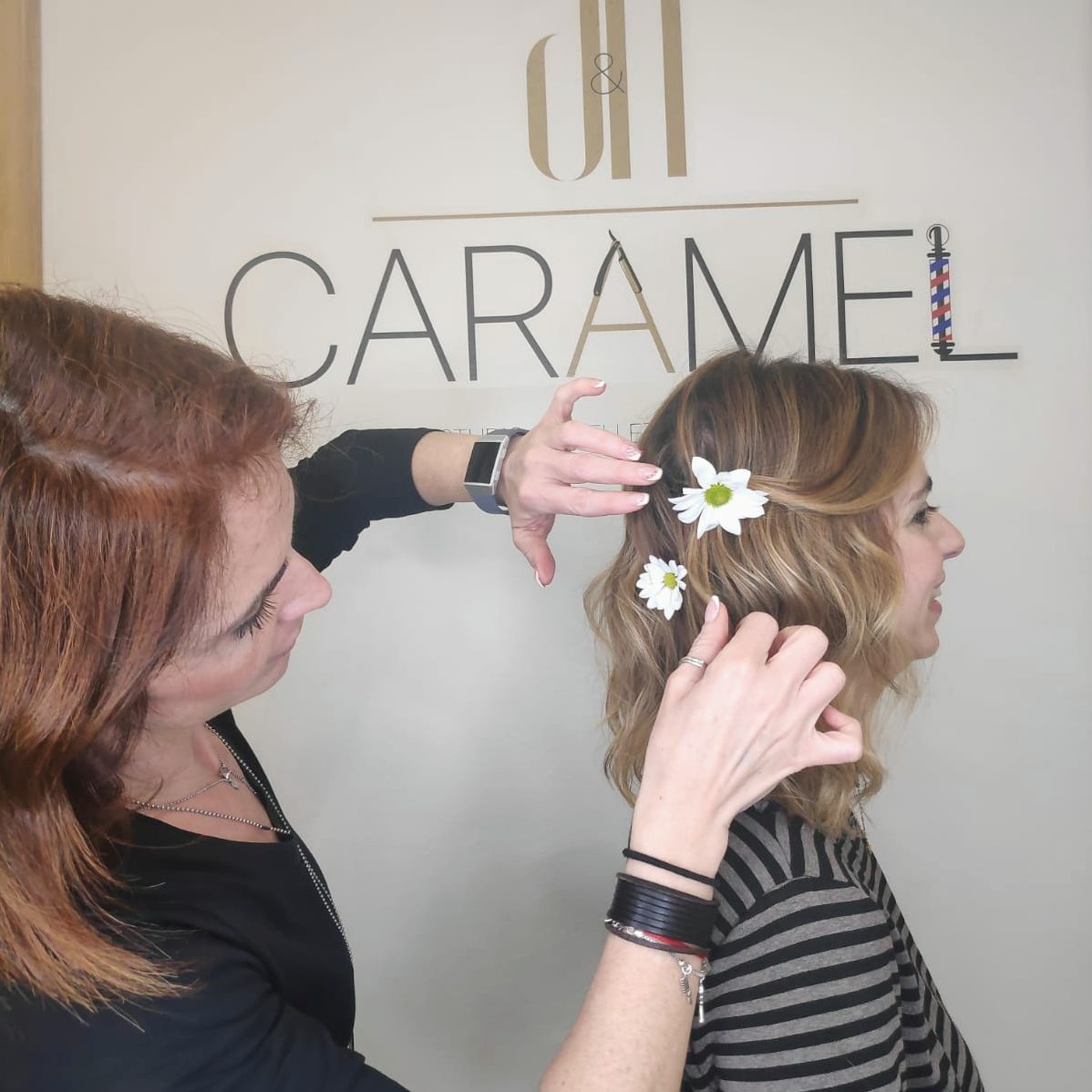 A stylist at A N CARAMEL adds floral accessories to a client's hair in a bright salon.