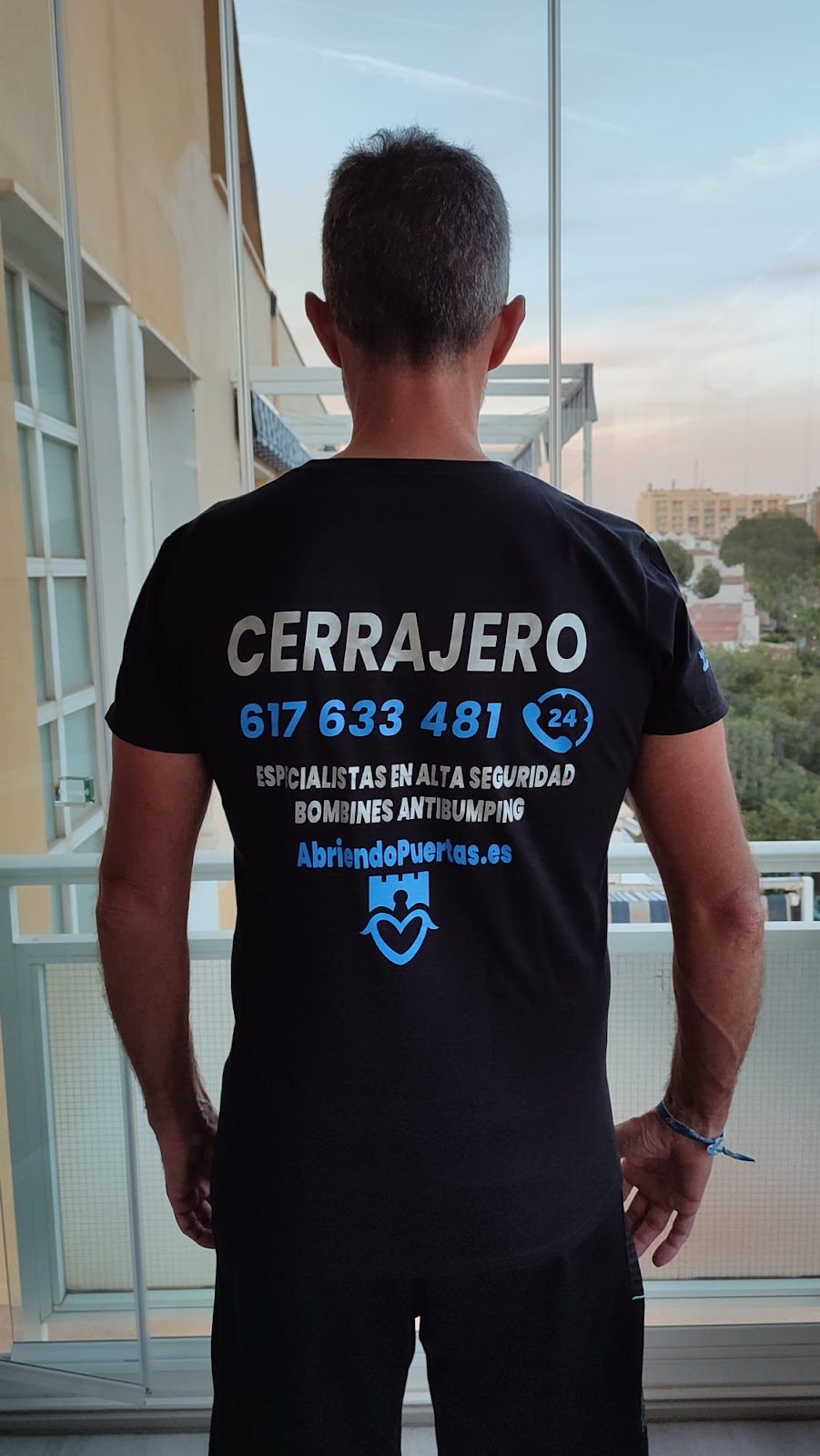 Una persona que viste una camiseta negra con la palabra "CERRAJERO" e información de contacto se encuentra en el interior de un edificio con vista a la ciudad.