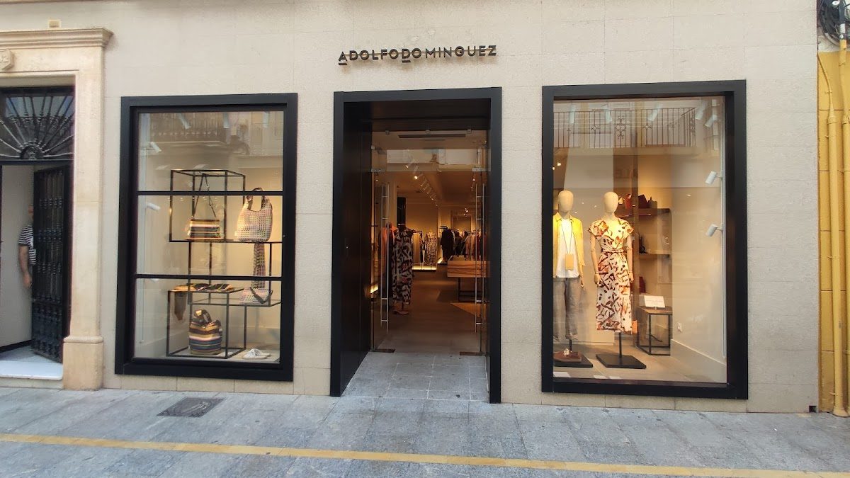 La tienda Adolfo Domínguez presenta elegantes exhibiciones de ropa en grandes escaparates de cristal en la Carrera Espinel.