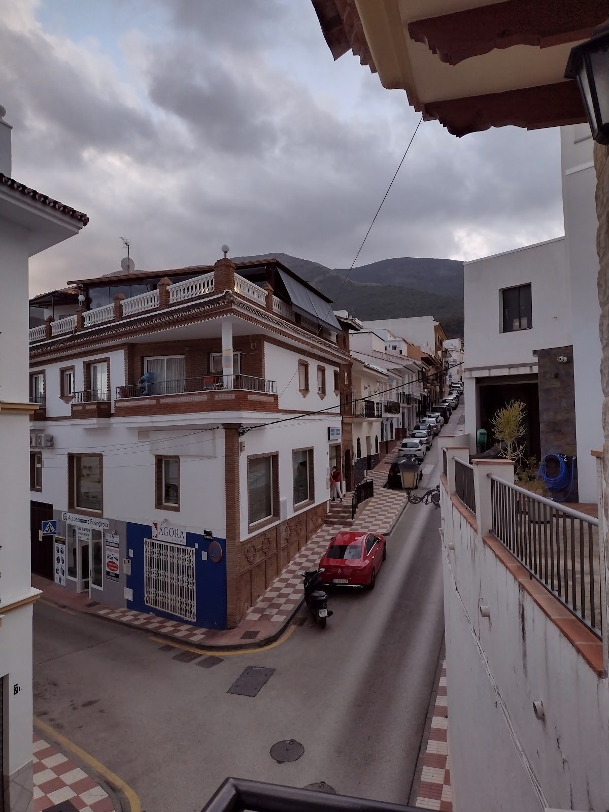 Vista exterior del Ágora Academia en una esquina de Alhaurín el Grande, con edificios cercanos y un coche rojo.