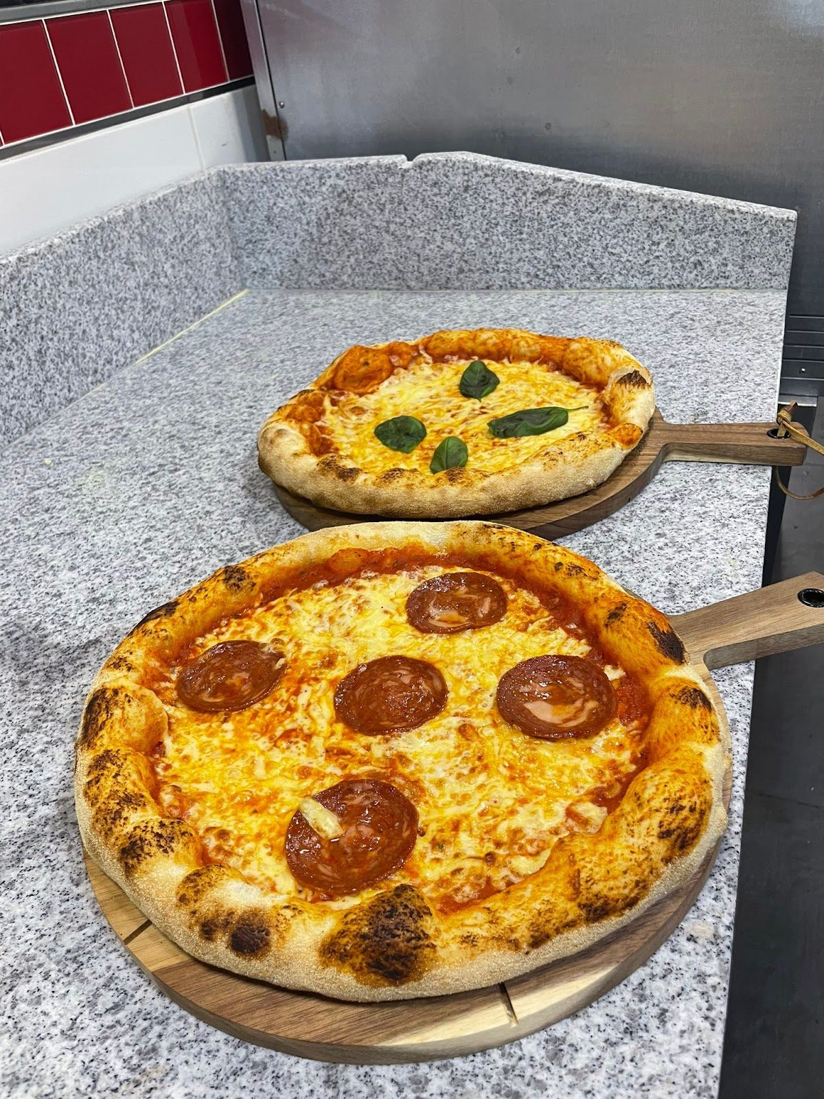 Dos pizzas recién horneadas sobre palas de madera, una con pepperoni y la otra con albahaca.