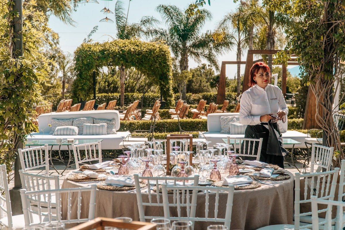 En Alabardero Catering & Experiences, en Málaga, se puede disfrutar de un espacio al aire libre con mesas elegantes, un miembro del personal y una exuberante vegetación.