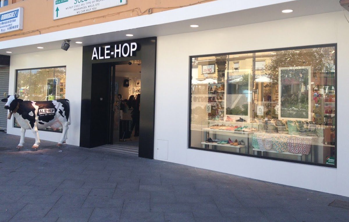 La tienda ALE-HOP presenta un exterior moderno con grandes ventanales que exhiben mercancía colorida y una estatua de una vaca en el exterior.