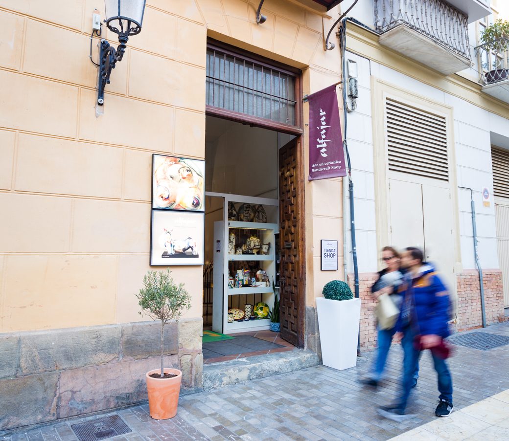 The entrance of ALFAJAR Cerámica Artesanal features a wooden door and displays artisanal ceramics inside.
