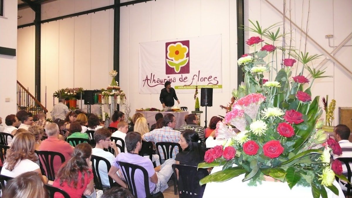 Un arreglo floral con los asistentes sentados, y un gran letrero de Alhaurina de Flores S.L. al fondo.