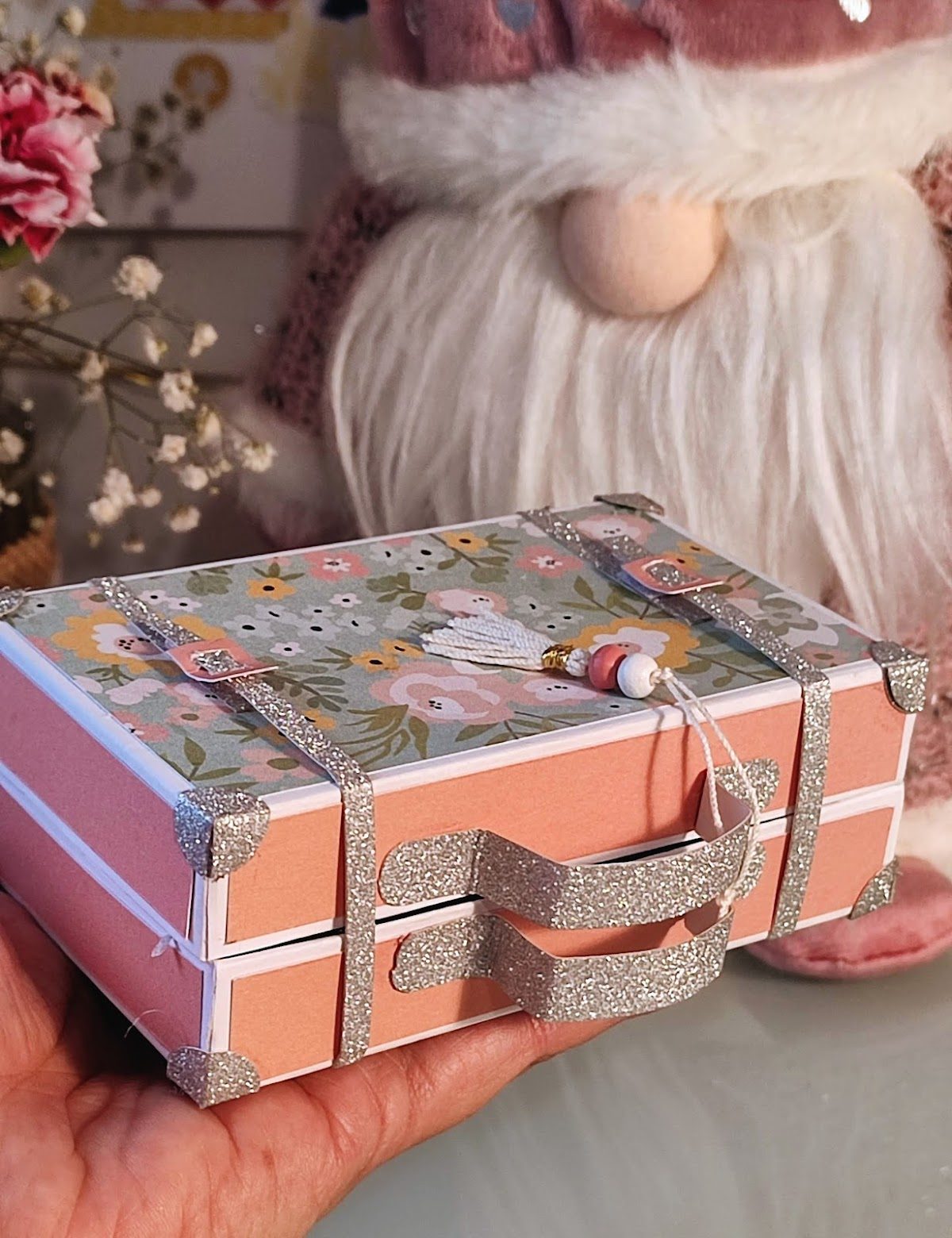 Una maleta miniatura decorativa con diseño floral, sostenida en una mano, rodeada de flores y una figura de peluche.