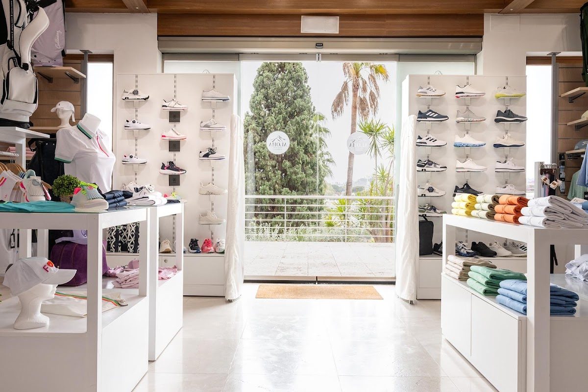 Vista interior de la tienda Aloha Golf Pro Shop, con expositores de ropa y zapatos, una entrada luminosa y zonas verdes en el exterior.