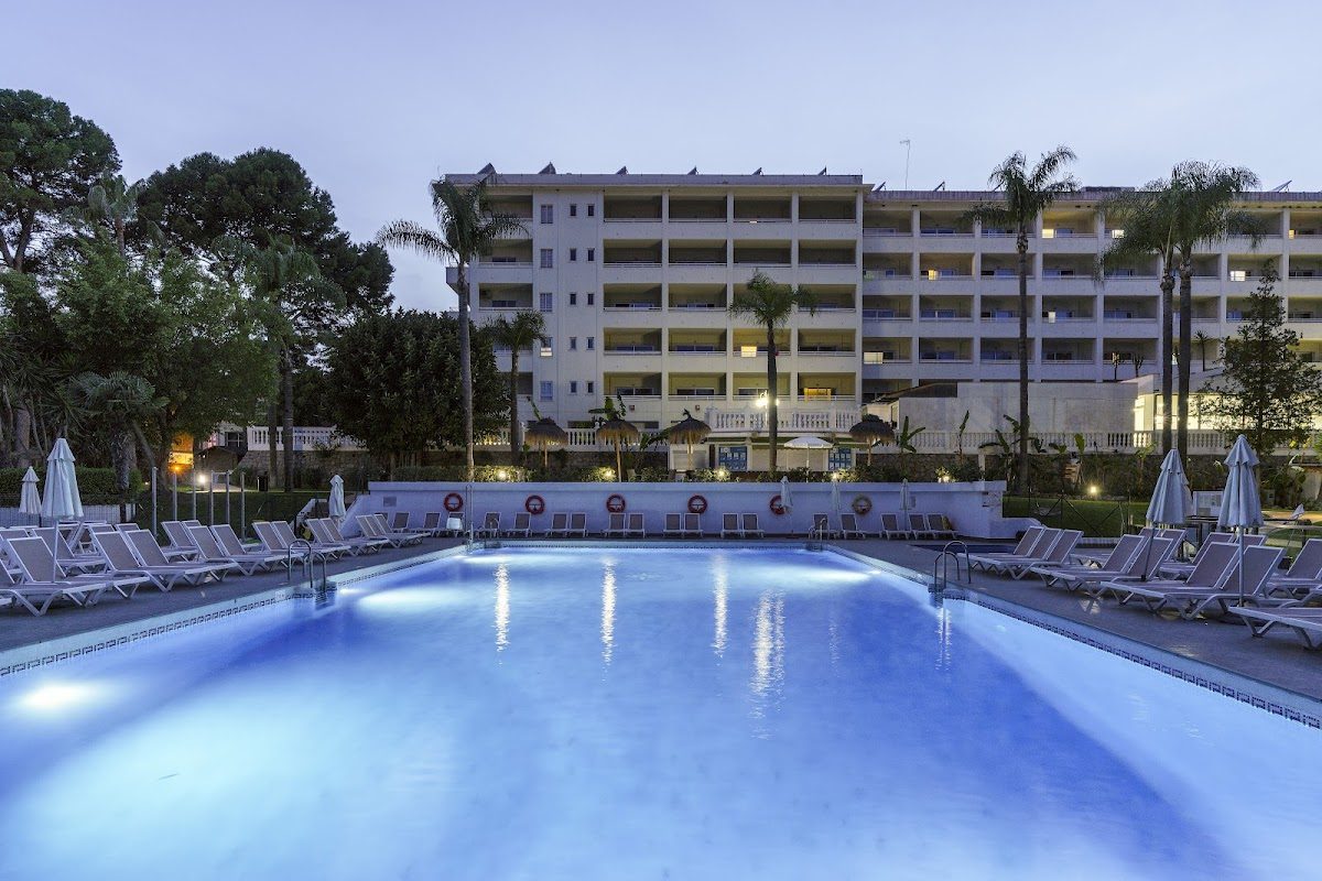La imagen muestra una zona de piscina bien iluminada en el Parque AluaSun Costa, rodeada de palmeras y tumbonas.
