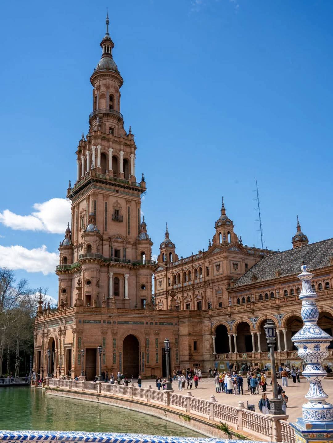 The image shows the stunning architecture of the Plaza de España in Seville, featuring intricate towers and a reflecting pool.