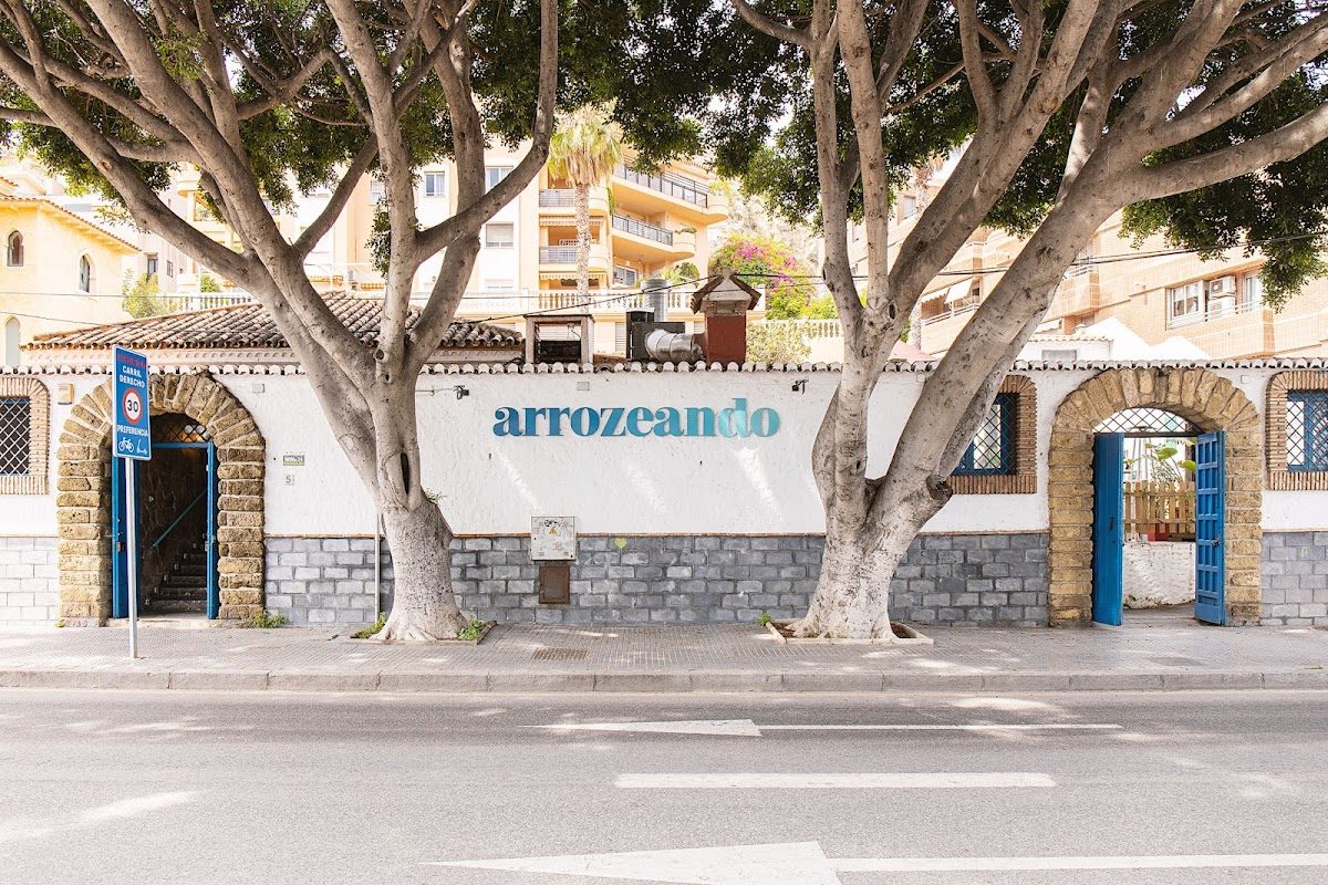 El exterior de Arrozeando Pedregalejo presenta una pared blanca con detalles azules y grandes árboles en la parte delantera.