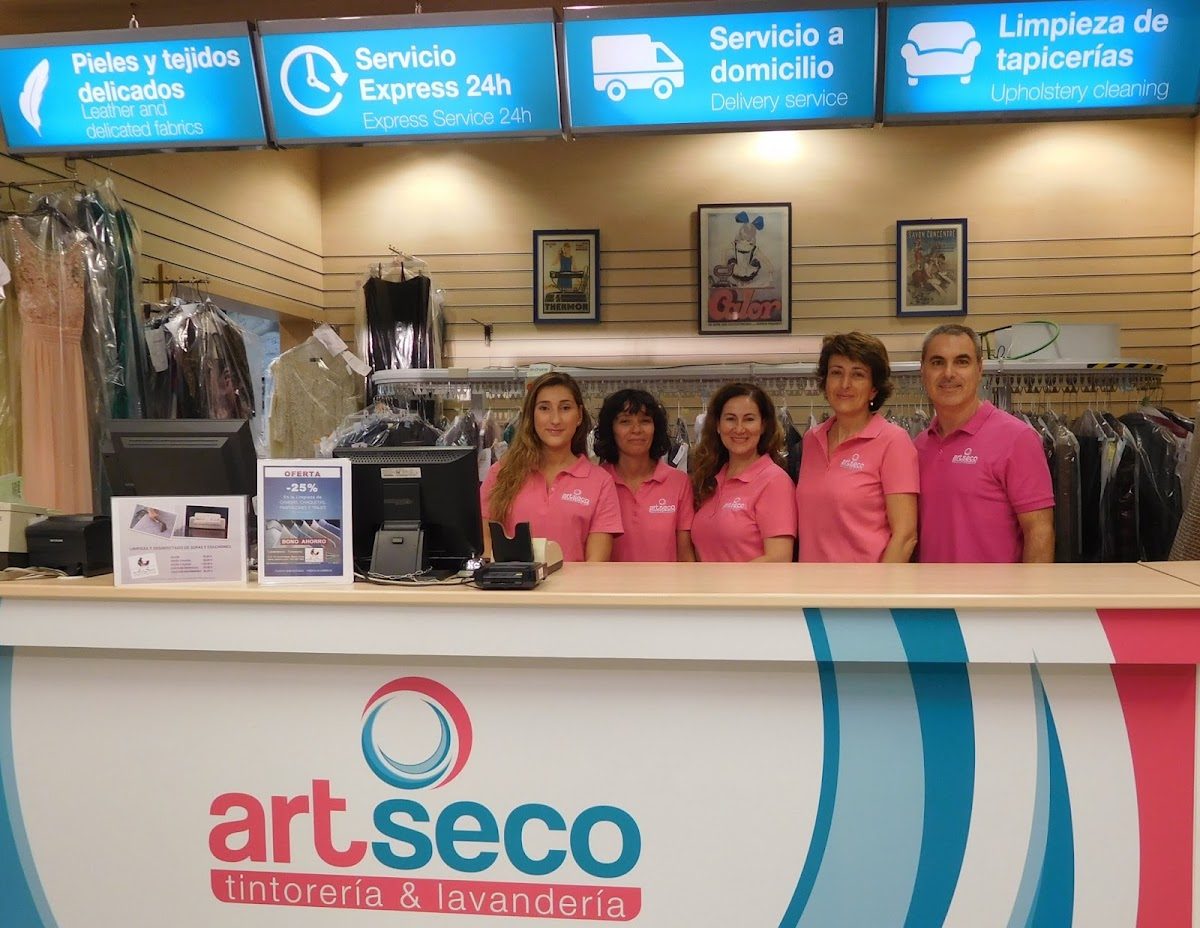 Personal de ARTSECO TINTORERIA LAVANDERIA con camisas rosas en la zona de recepción, con los servicios de limpieza expuestos detrás de ellos.