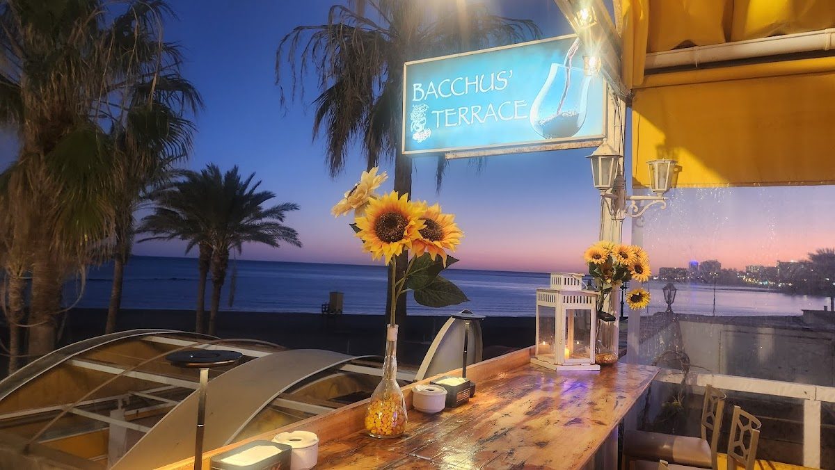 La terraza de Bacchus cuenta con una barra de madera decorada con girasoles y ofrece una hermosa vista de la playa al atardecer.