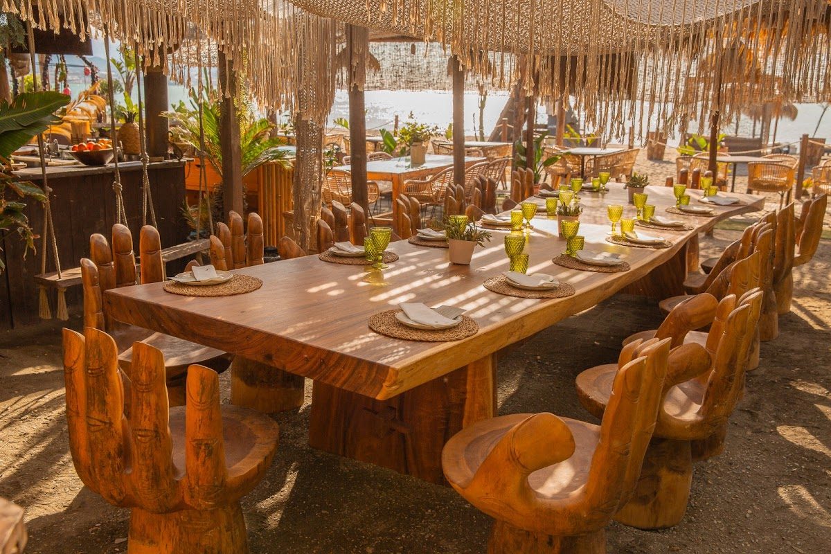 Zona de comedor al aire libre en Bahía Beach Jazba con mesas y sillas de madera, rodeada de plantas y con vistas a la playa.