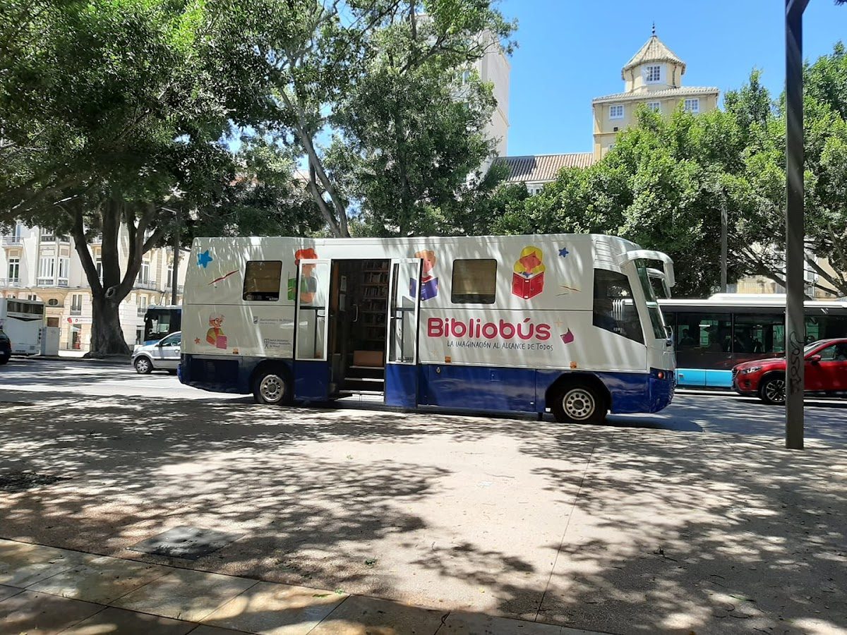 A mobile library bus with colorful graphics parked under trees on a city street.
