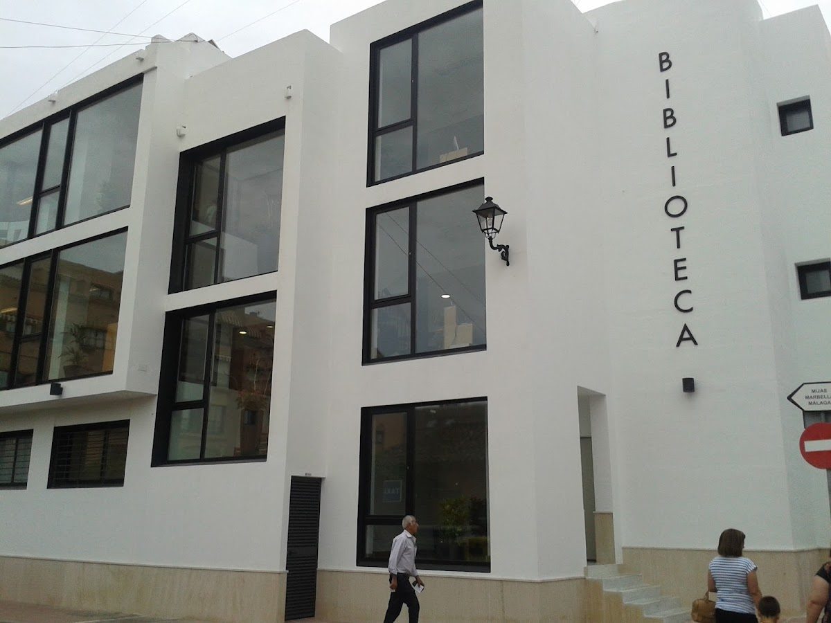 Modern white building with large windows, featuring the sign "BIBLIOTECA" and people walking outside.