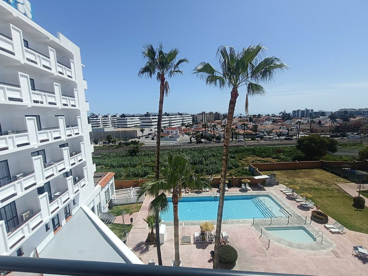 View from a balcony at BLUESEA Costa Serena, showcasing a pool surrounded by palm trees and the landscape beyond.