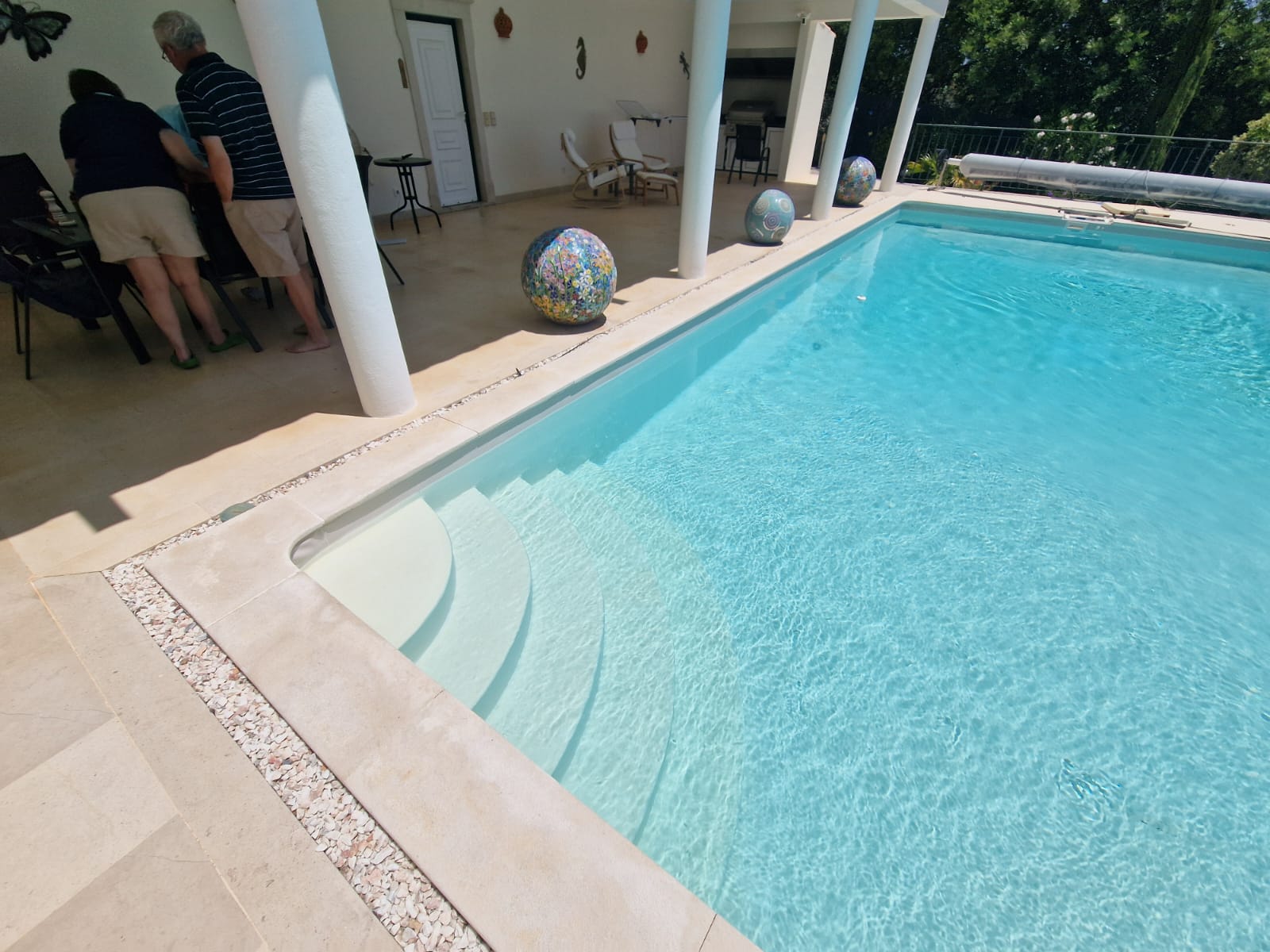 Una piscina cristalina con escalones, rodeada de asientos y esferas decorativas en una soleada zona al aire libre.