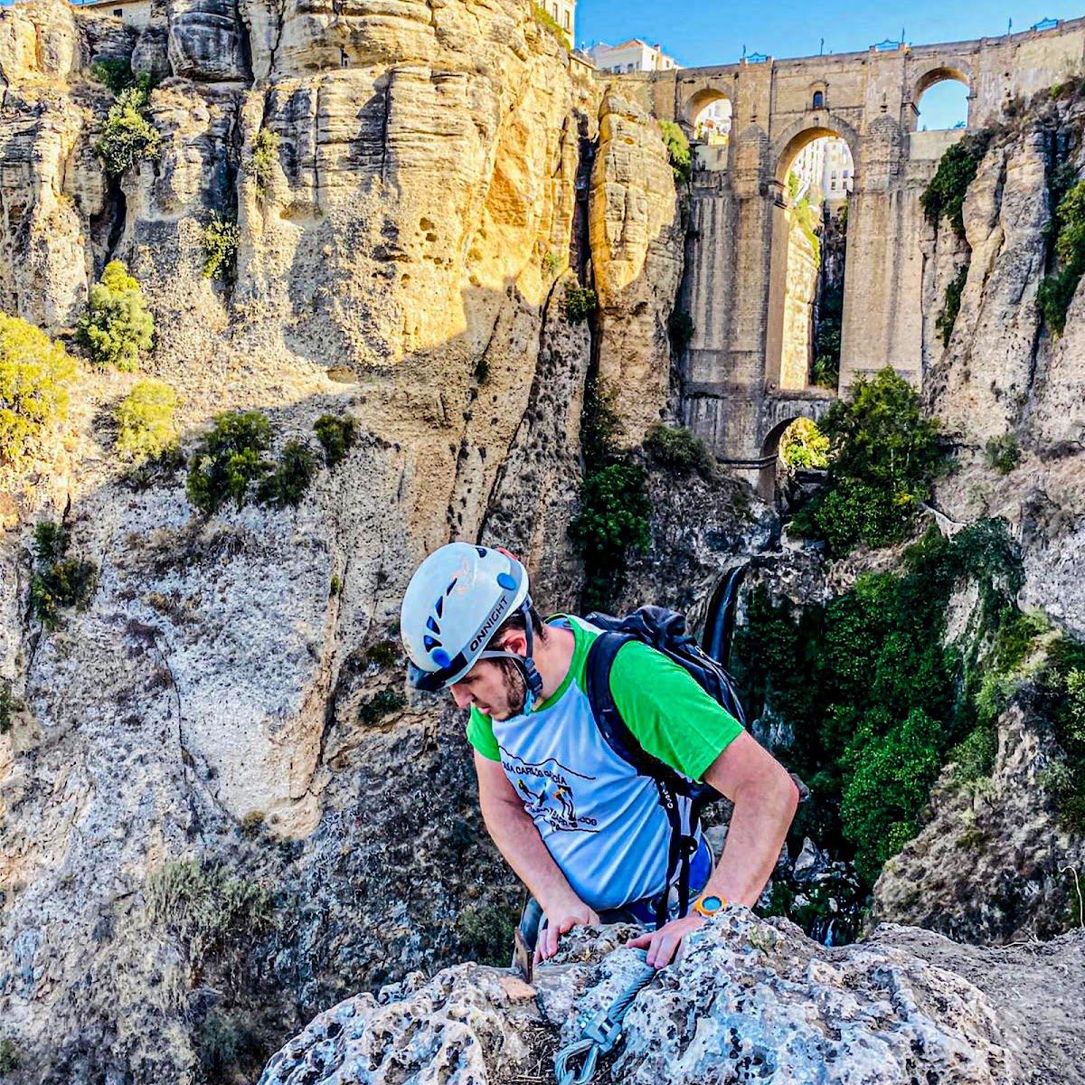 A climber in a helmet navigates rocky terrain with a bridge and waterfall in the background at Carlos J. - Guía de Montaña y Barrancos.