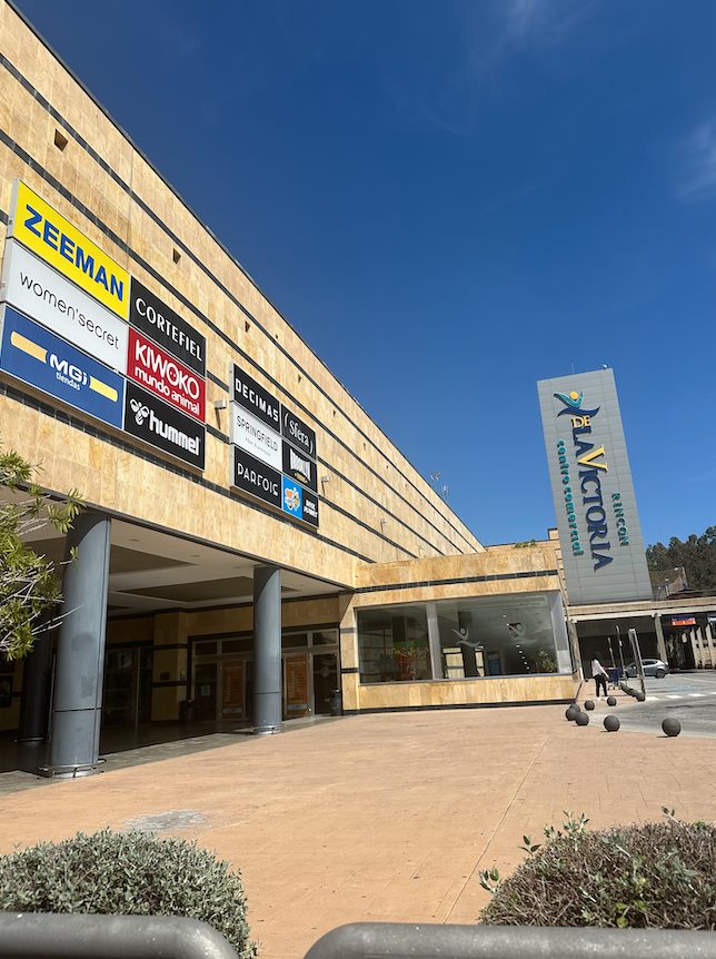 The exterior of Centro Comercial Rincón de la Victoria features various store signs and a clear blue sky.