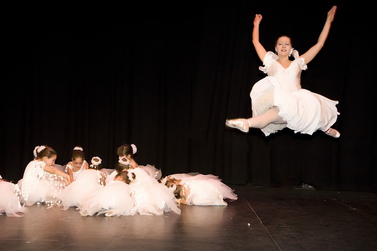 Una bailarina con un vestido blanco salta con gracia por encima de otras jóvenes bailarinas también con vestidos blancos, sentadas en el escenario.