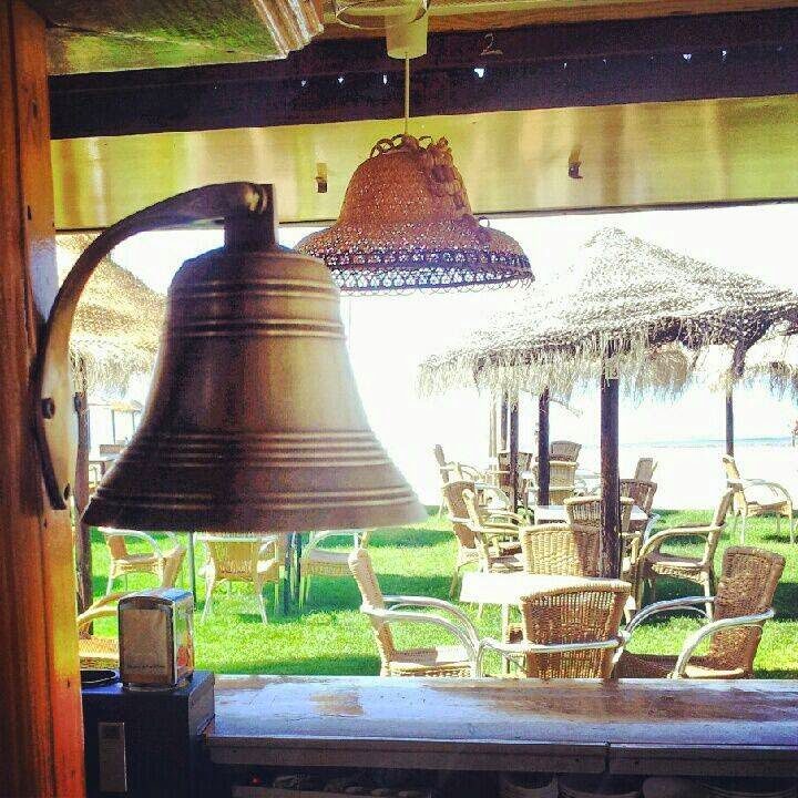 View from inside Chiringuito Cacola, featuring a bell and outdoor seating with straw umbrellas by the beach.