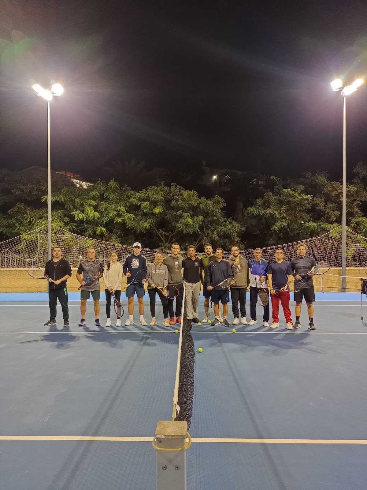 A group of tennis players stands on a blue court at Club de Tenis Sohail, illuminated by floodlights at night.