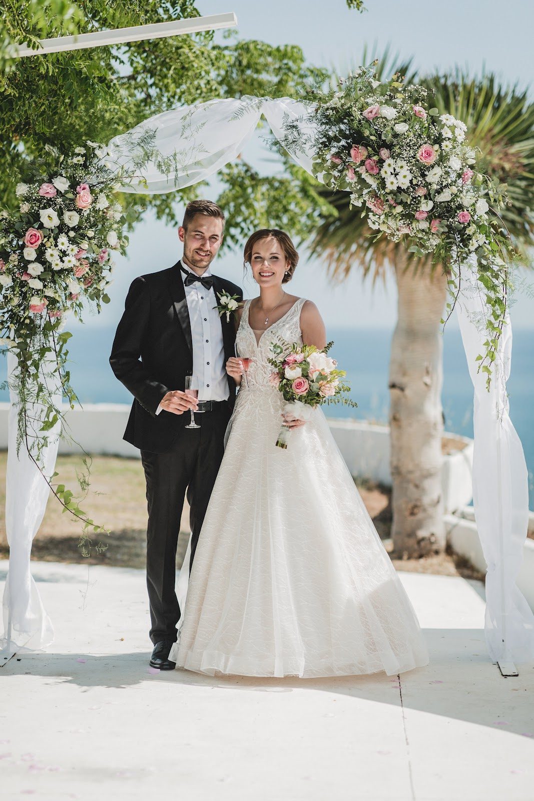 Una pareja de recién casados posa bajo un arco floral, sonriendo con un paisaje pintoresco de fondo.