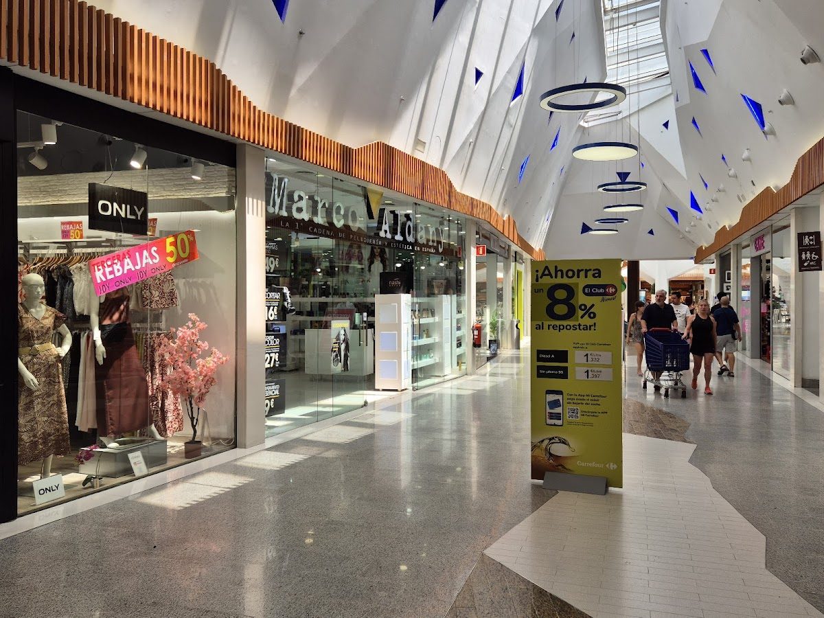 Vista interior del centro comercial Costasol, con tiendas, compradores y luces decorativas en el techo.