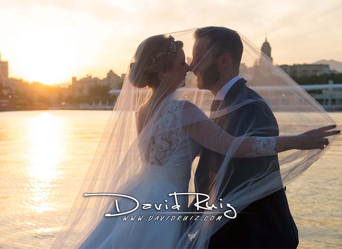 Una pareja se abraza junto al agua al atardecer, con un velo que crea una atmósfera romántica para su boda.