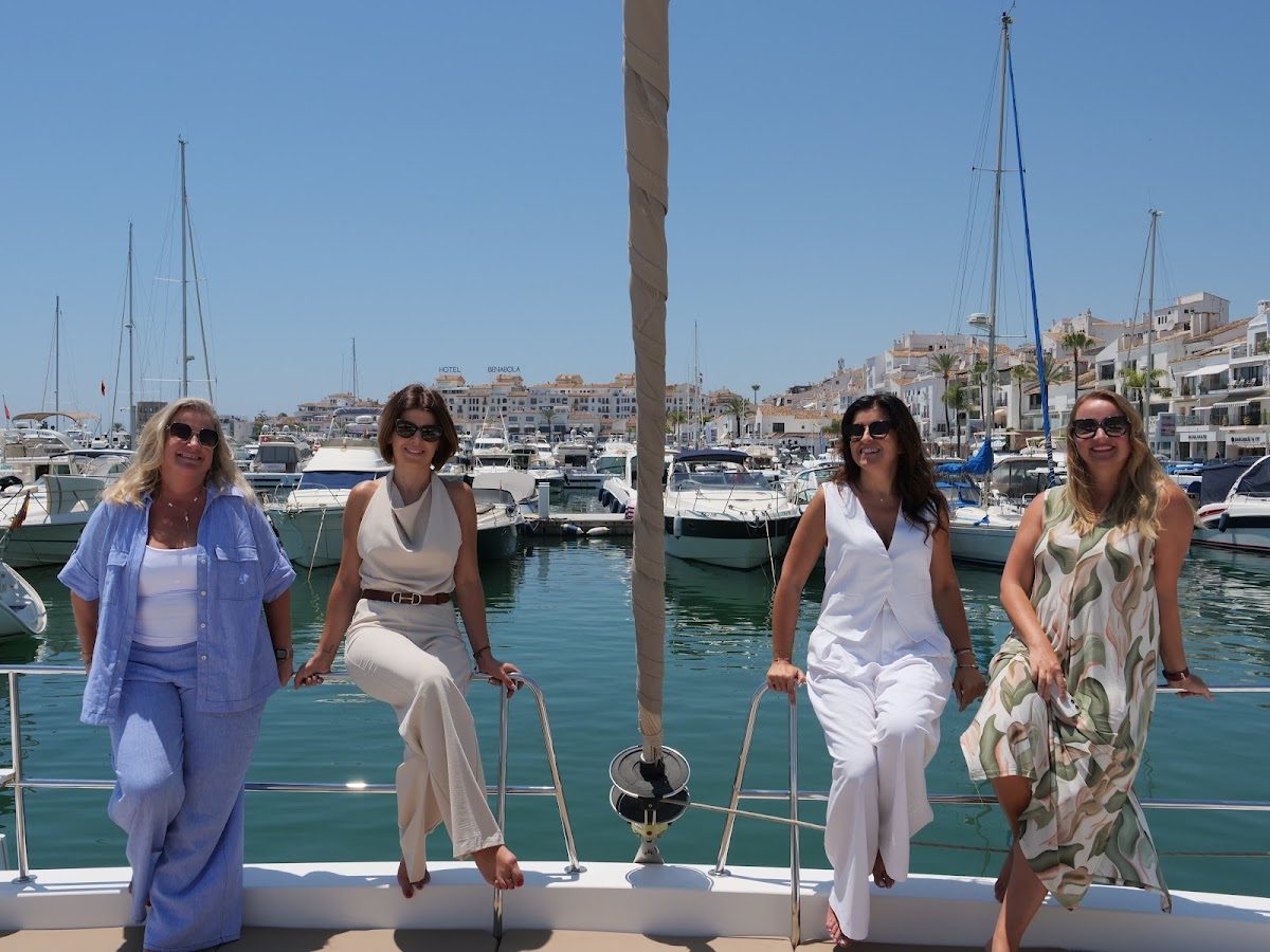 Cuatro mujeres posan en un barco en un puerto deportivo, rodeadas de yates y con un pueblo costero al fondo.