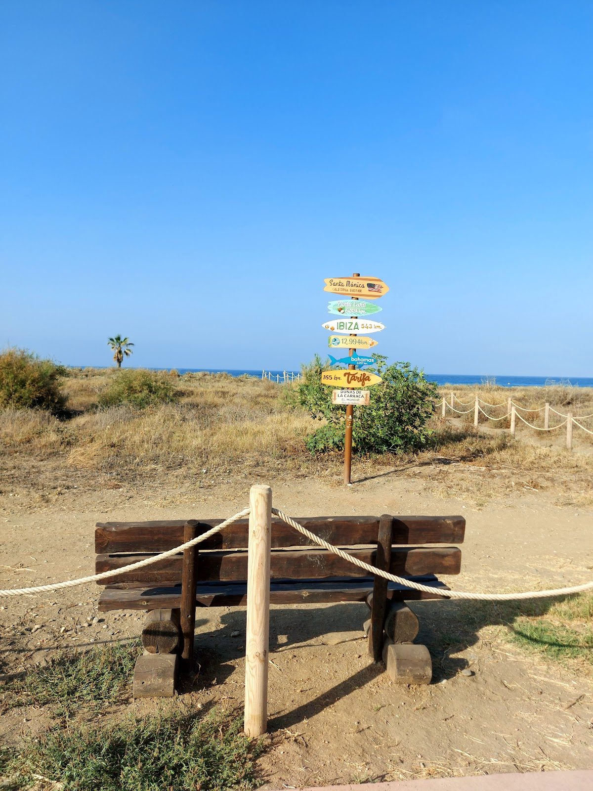 A wooden bench with directional signs pointing to various destinations near the coastal dunes of Dunas de La Carraca.