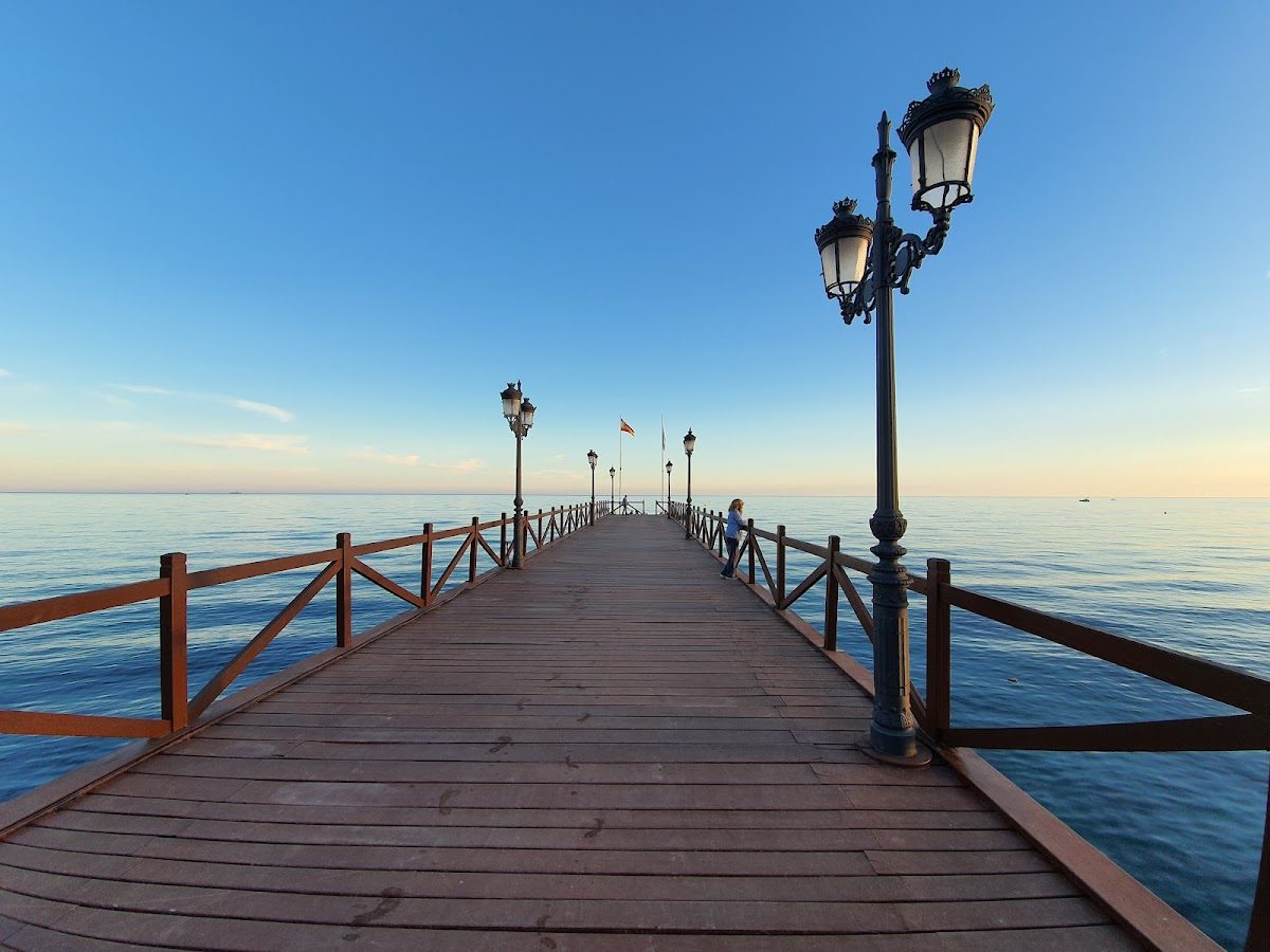 Un muelle de madera se extiende sobre aguas tranquilas, con faroles decorativos y banderas ondeando a lo lejos.