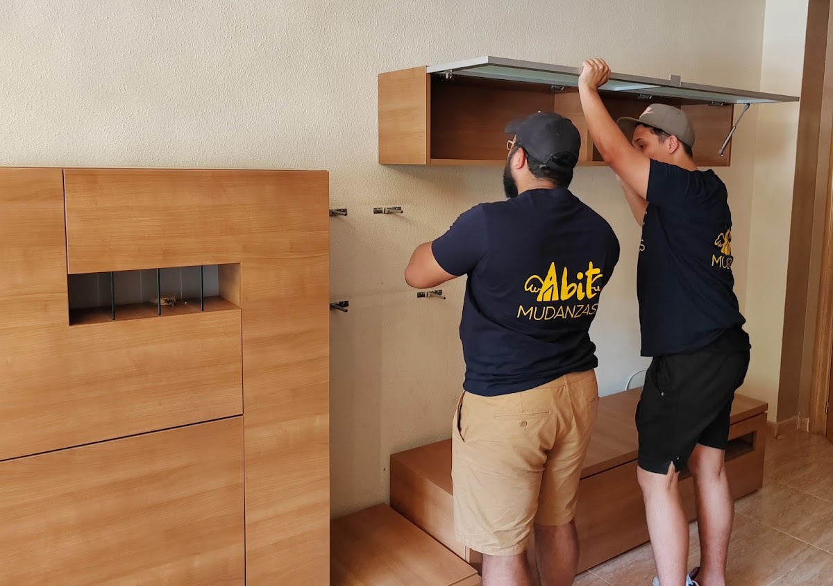 Two workers from Abit Mudanzas are assembling furniture in a modern interior space.