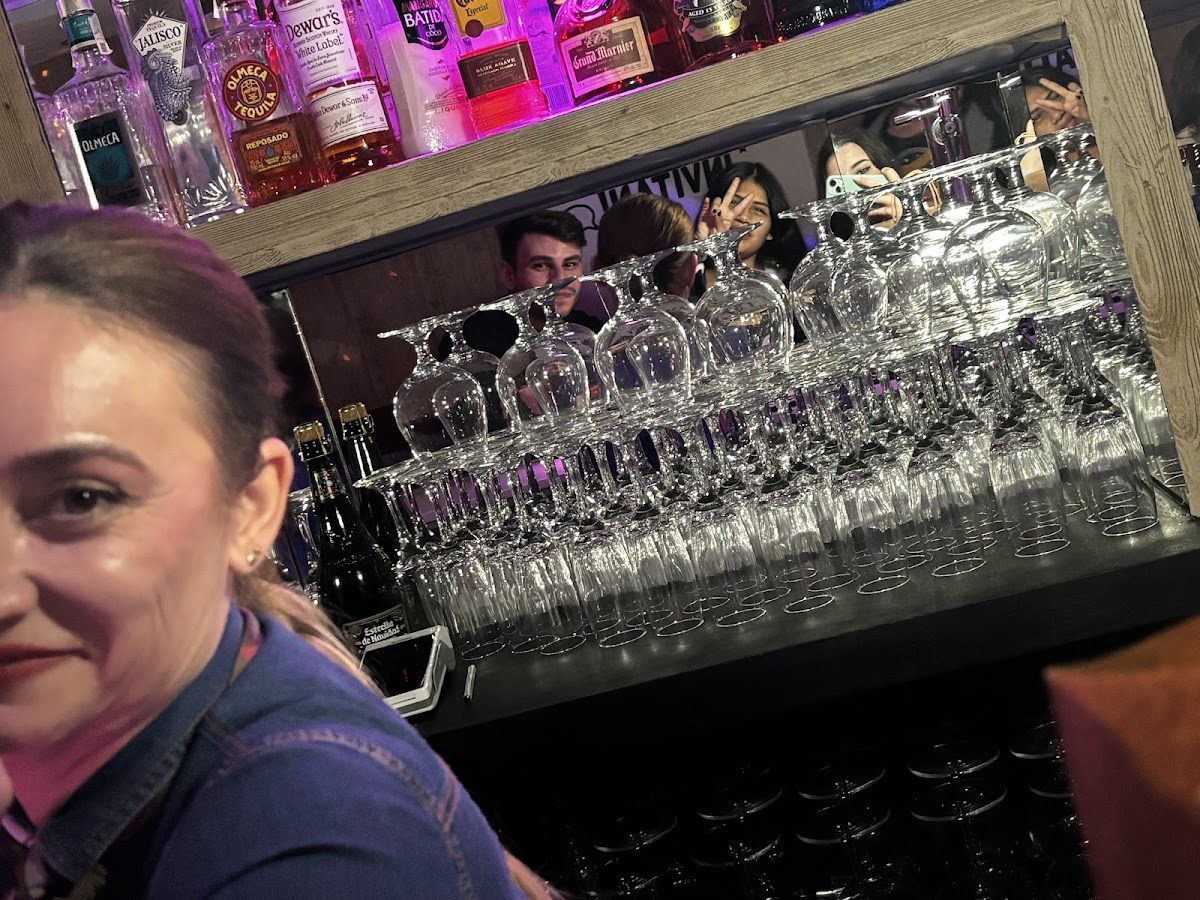 A bartender stands in front of a shelf filled with glassware and colorful liquor bottles at Esencia.