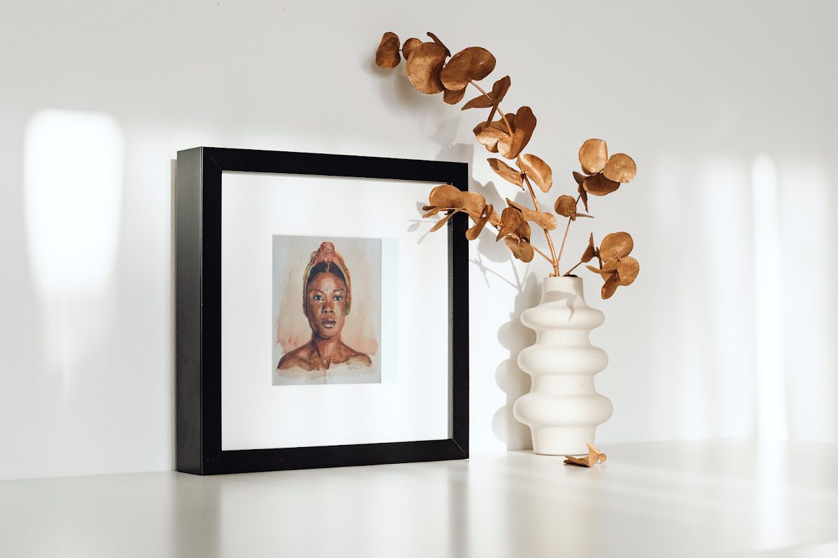 A framed portrait of a woman next to a decorative vase with dried flowers on a white shelf.