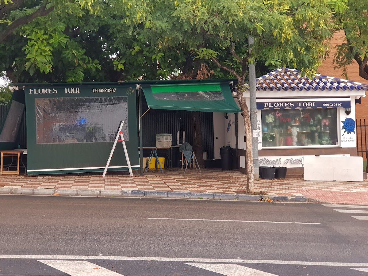 Flores Tobi presenta un exterior verde con señalización, rodeado de árboles y una acera de baldosas en San Pedro Alcántara.