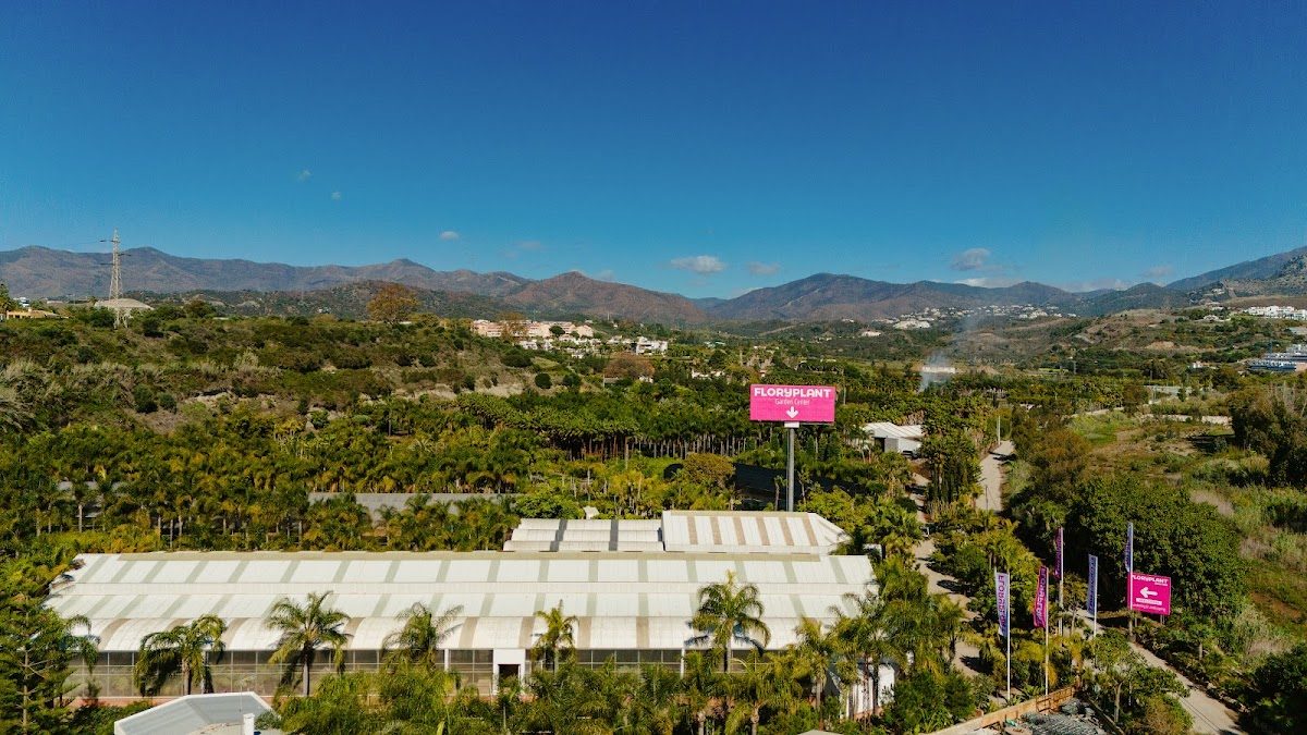 El centro de jardinería FLORYPLANT cuenta con un gran invernadero rodeado de exuberante vegetación y montañas al fondo.