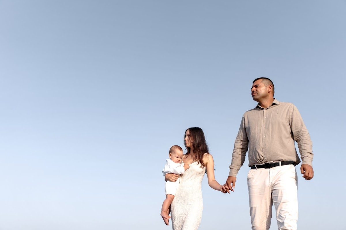 A family portrait featuring a woman holding a baby and a man standing beside her against a clear blue sky.