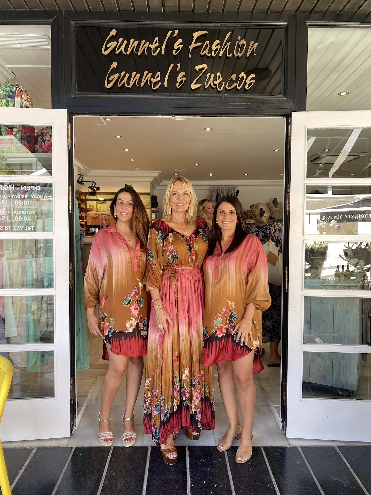 Three women stand outside Gunnel's Fashion Marbella, showcasing colorful dresses in a stylish boutique entrance.