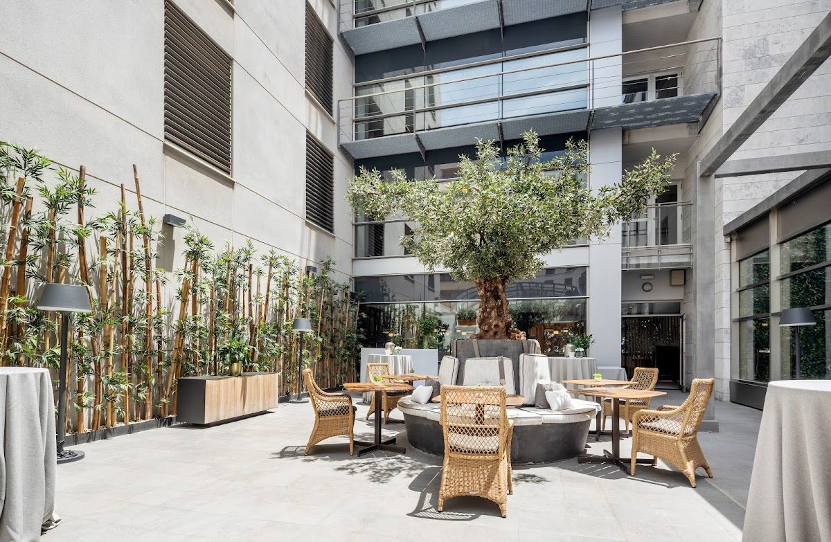 Comedor al aire libre en el Hotel ILUNION Málaga con sillas de mimbre, un árbol central y decoración de bambú.