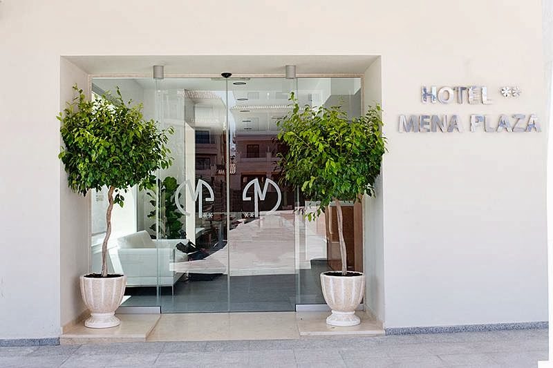 The entrance of Hotel Mena Plaza features glass doors and potted trees on either side.