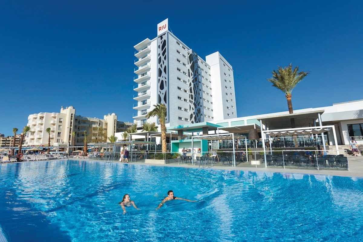 Hotel Riu Costa del Sol features a large outdoor pool with guests swimming and palm trees in the background.