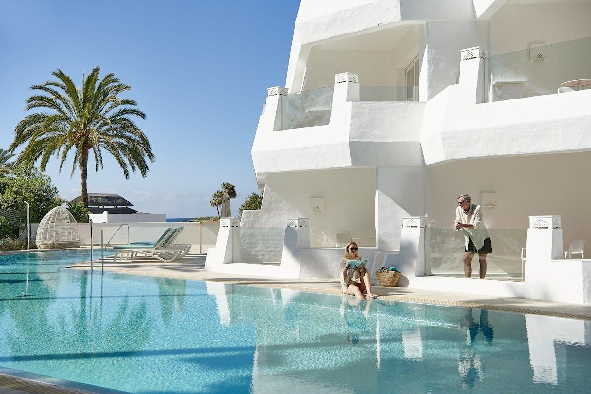La imagen muestra una moderna zona de piscina con edificios blancos, palmeras y huéspedes relajándose junto al agua.