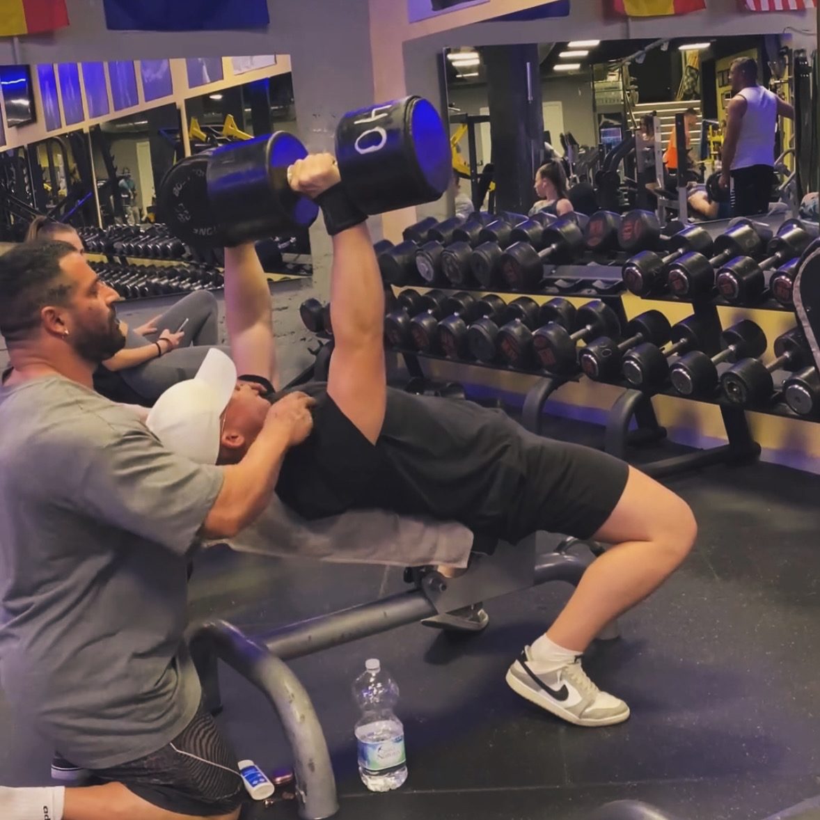 Un entrenador ayuda a un hombre a levantar pesas en un banco en la zona de entrenamiento del gimnasio Iron Gym.