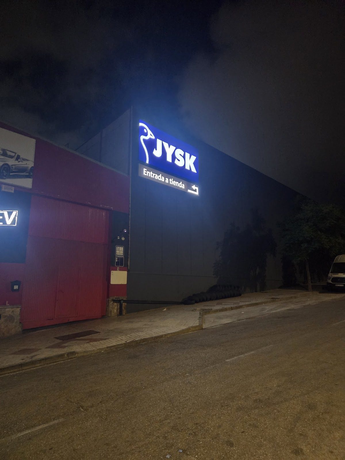 El letrero iluminado de JYSK por la noche, que indica la entrada a la tienda, con un edificio oscuro y vista de la calle.