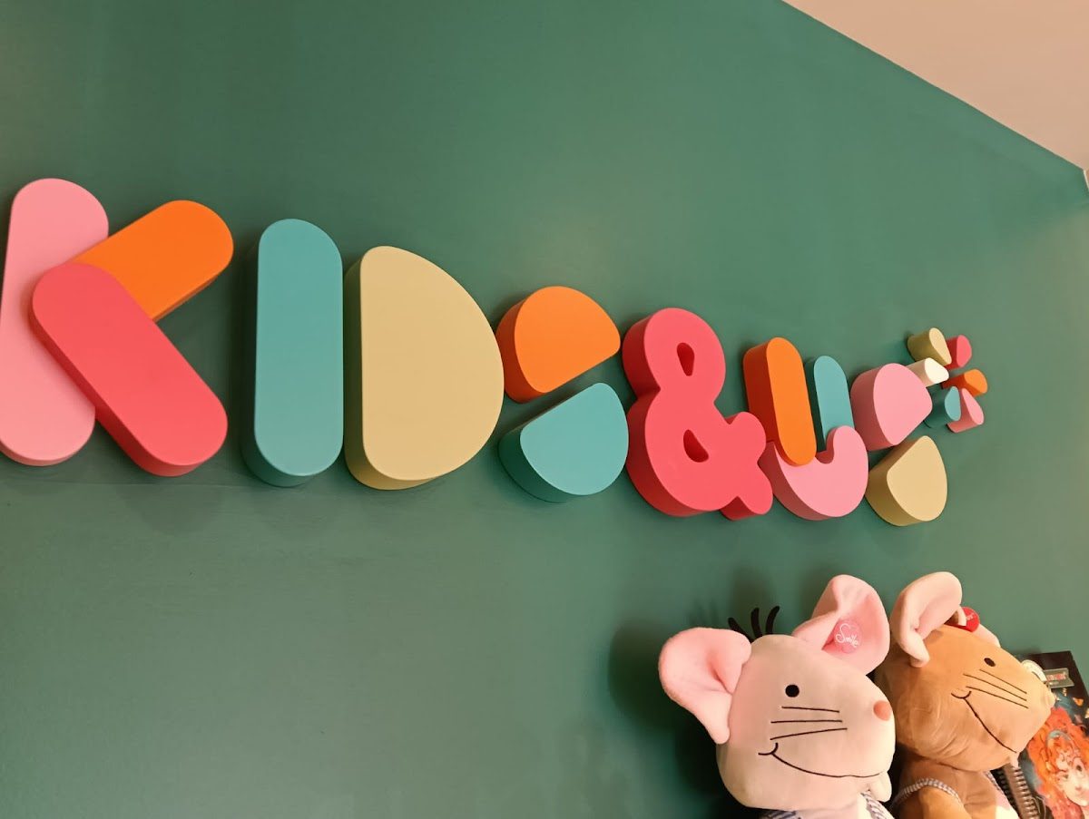 Letrero colorido con la leyenda "Kids&Us" y juguetes de peluche en una pared verde de la academia de Benalmádena.